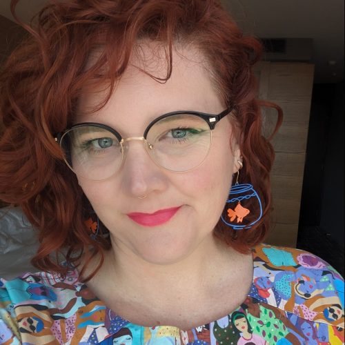 An image of a woman with red hair and glasses. She is wearing a colourful dress and earrings with fish.